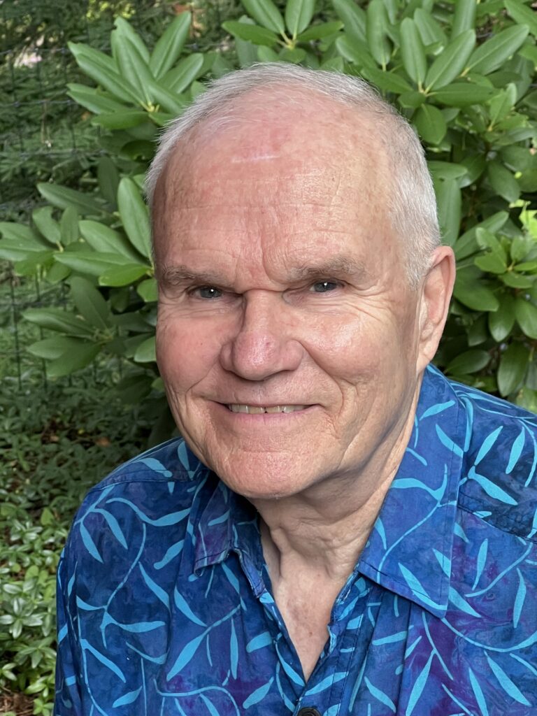 An older man with short white hair wearing a blue patterned shirt smiles in front of lush green foliage.