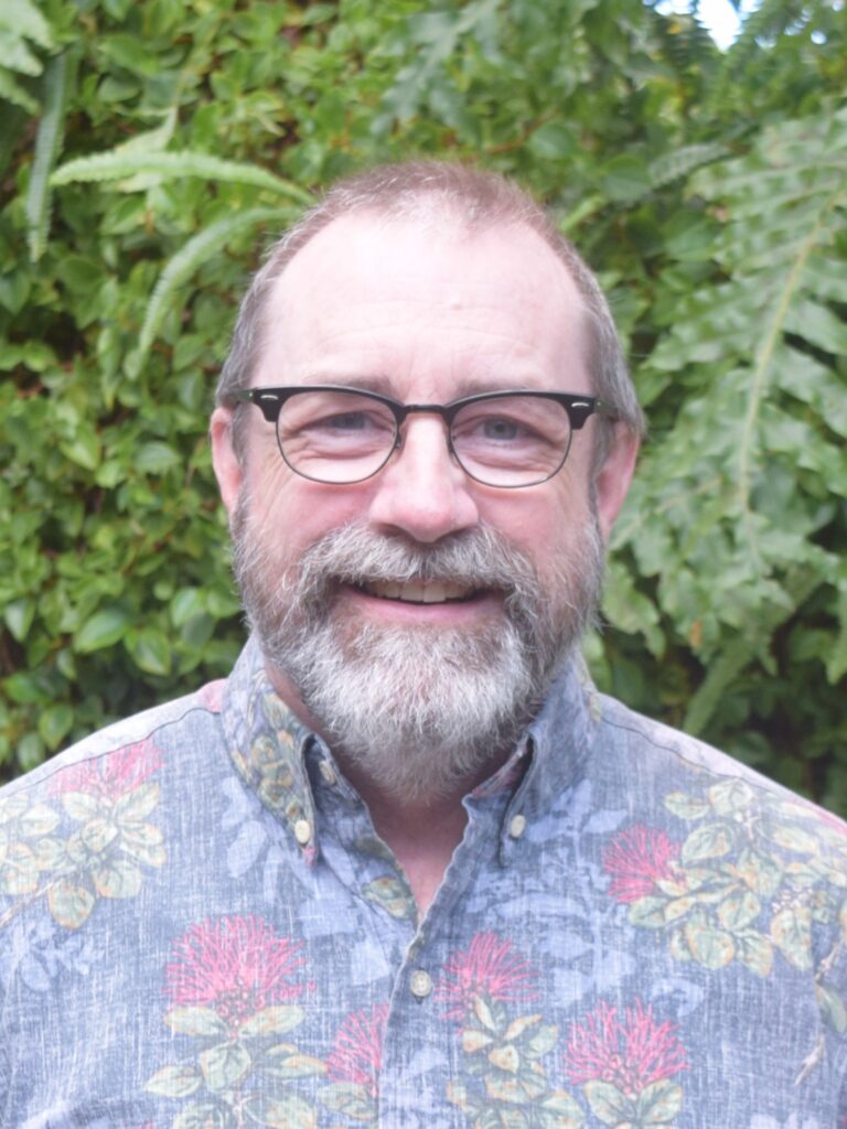 A middle-aged man with glasses and a gray beard smiles at the camera, standing outdoors in front of green foliage.